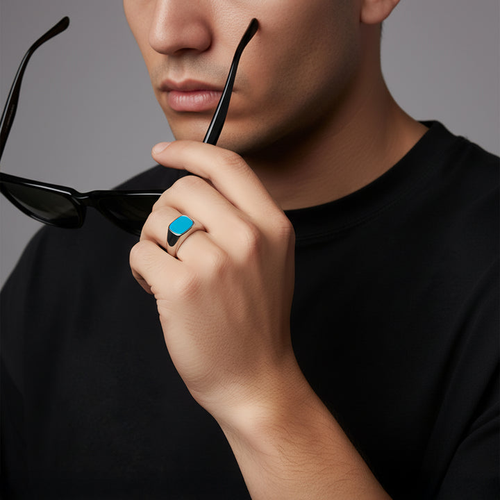 Man wearing black sunglasses and a black shirt with a blue stone signet ring on his finger against a gray background